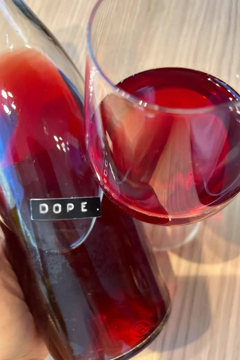 A close-up of a hand holding a glass bottle labeled 'DOPE.' next to a stemmed wine glass filled with deep rosé wine, set on a light wood surface. Preisinger Dope - Natural Wine Dealers