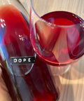 A close-up of a hand holding a glass bottle labeled 'DOPE.' next to a stemmed wine glass filled with deep rosé wine, set on a light wood surface. Preisinger Dope - Natural Wine Dealers